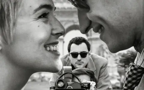Primo piano di una coppia che ride, con un uomo che filma sullo sfondo, evocando l'estetica dei registi della nouvelle vague.