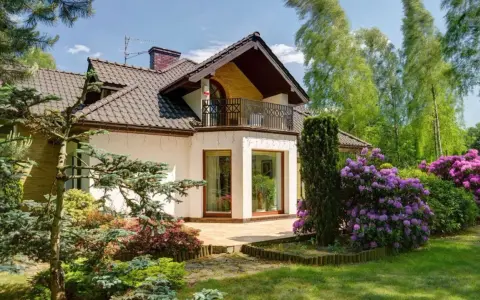 Dom z balkonem i tarasem, otoczony bujną zielenią i kwitnącymi rododendronami. Występ ściany podkreśla nowoczesny charakter budynku.