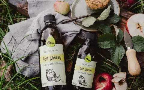 Dwie butelki naturalnego octu jabłkowego Olini, idealnego dla dzieci, leżą wśród jabłek i liści.