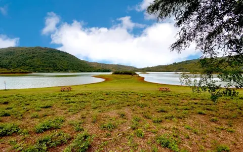 Prato verde con tavoli da picnic affacciati sul **lago di Canterno**, circondato da dolci colline sotto un cielo azzurro con nuvole bianche.