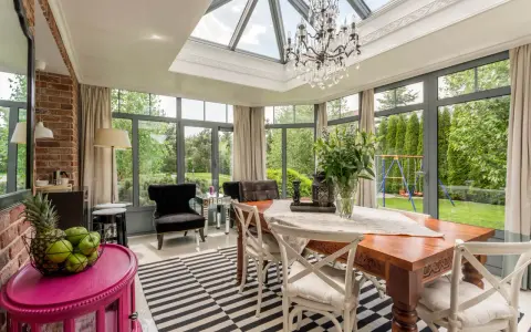 Une élégante veranda couloir avec un lustre, une table en bois et des fauteuils, donnant sur un jardin verdoyant.