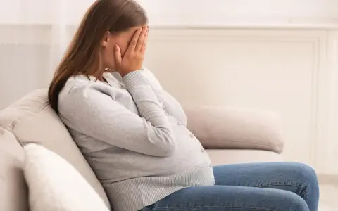 Kobieta w ciąży siedzi na kanapie, zakrywając twarz dłońmi. Jej postawa wyraża głęboki smutek, być może związany z diagnozą wady letalnej.
