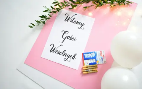 Tablica powitalna wesele z napisem "Witamy Gości Weselnych", ozdobiona gałązką zieleni i lampkami. Obok taśma, złote pisaki i białe balony.