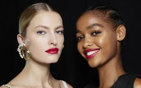 Dos modelos lucen un maquillaje para boda con labios rojos vibrantes y piel radiante. Una lleva un pendiente de perlas, la otra sonríe.