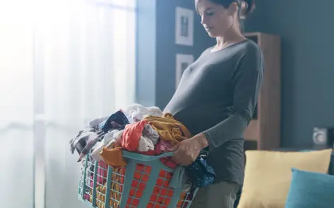 Ciężarna kobieta z koszem prania. Dźwiganie w ciąży bywa wyzwaniem, ale ona radzi sobie z codziennymi obowiązkami.