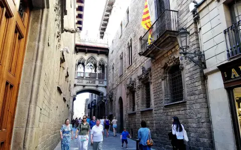 Menschen schlendern durch eine enge Gasse in der **Barcelona Altstadt**. Über ihnen spannt sich eine historische Brücke mit filigranen Bögen.