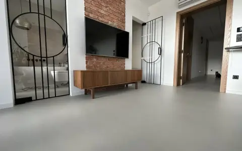 Nowoczesne wnętrze z betonową podłogą, co to jest mikrocement, która nadaje mu surowego charakteru. W tle widać łazienkę i salon.