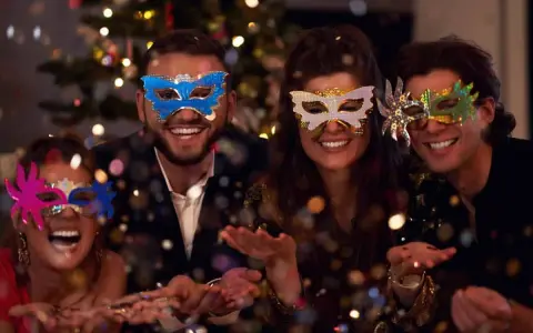 Cuatro amigos disfrutan de una temática fiesta de mascaras, riendo y lanzando confeti frente a un árbol de Navidad iluminado.