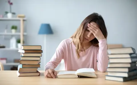 Zmęczona dziewczyna z ręką na czole przy stosach książek, próbująca napisać wypracowanie.