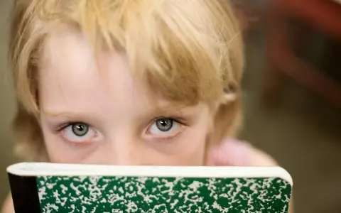 Oczy dziecka z blond włosami spoglądają zza zielonego zeszytu, ukrywając niepewność związaną z fobią szkolną.