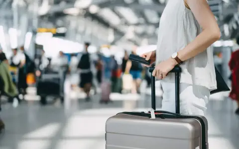 A traveler holds a passport and suitcase, mindful of carry-on size limits in a busy airport.