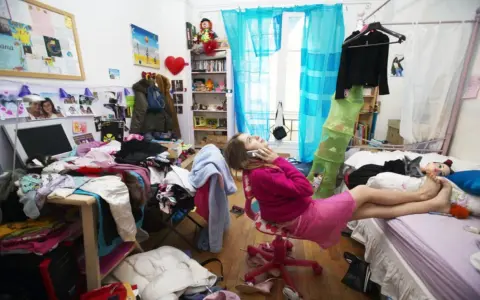 Chica en su habitación desordenada, hablando por teléfono, rodeada de ropa y objetos esparcidos.