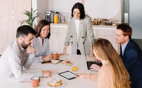 Equipo colaborando en una mesa con café y bocadillos. A pesar de las reuniones largas, el ambiente es positivo y productivo.