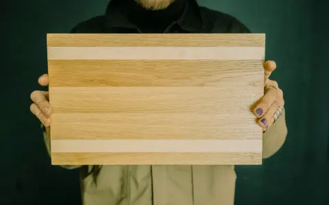 Hände halten ein rechteckiges Schneidebrett aus naturbelassenen holzbrettern mit hellen und dunkleren Streifen.