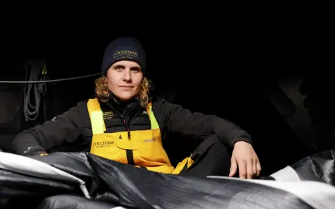 Clarisse Crémer, Seglerin im L'Occitane Sailing Team, sitzt in dunkler Kleidung und gelber Latzhose inmitten von Segeln.
