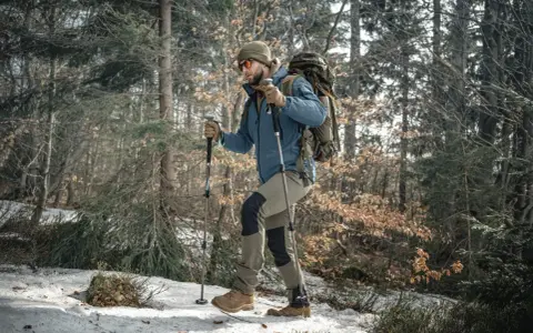 Mężczyzna w zimowej kurtce i czapce idzie przez las, wspierając się na kijach trekkingowych. Śnieg i suche liście pokrywają ścieżkę.