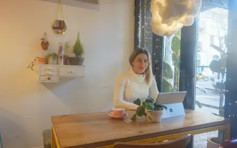Kobieta pracuje na laptopie w kawiarni, ciesząc się kawą i roślinami. To idealne miejsce na dodatkową pracę zdalną.