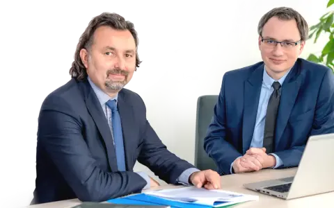 Dwóch mężczyzn w garniturach siedzi przy biurku z dokumentami i laptopem.