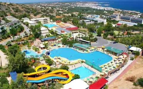 Widok z lotu ptaka na kompleks basenów z atrakcjami wodnymi i leżakami. Wiele osób korzysta z uroków słońca i wody. W tle widać budynki hotelowe i morze. Aqua Sun Village opinie potwierdzają, że to idealne miejsce na wakacje.