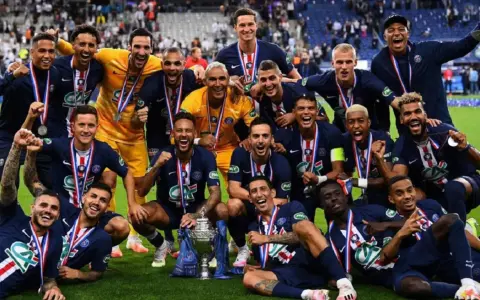 Un grupo de jugadores de Ligue 1 celebra con trofeos y medallas, sonriendo y vitoreando en el campo.