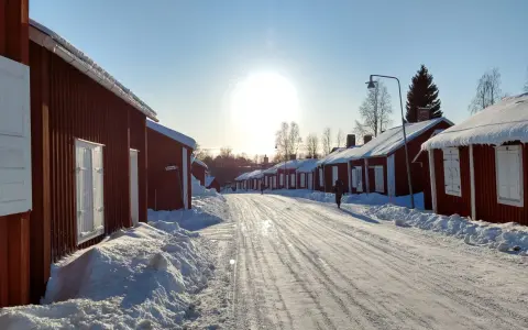 Vinterdag i kyrkbyn Gammelstad. Röda trähus med snö på taken kantar en snötäckt väg. Solen skiner starkt på en klarblå himmel.