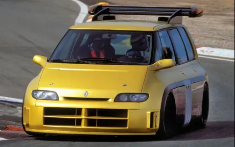 Żółty Renault Espace F1 z kierowcą w kasku na torze wyścigowym. Potężny spojler dachowy i agresywny wygląd.