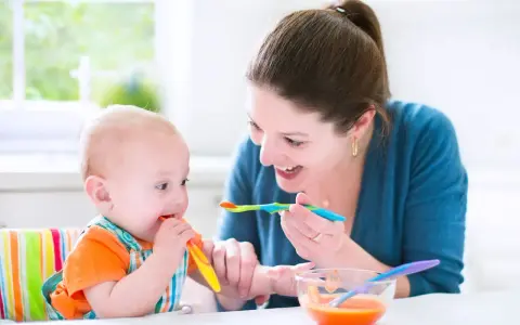 Uśmiechnięta mama karmi łyżeczką 7 miesięczne dziecko, które samo próbuje jeść.