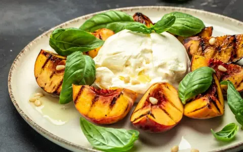 Burrata z grillowanymi brzoskwiniami, bazylią i orzeszkami piniowymi na talerzu.