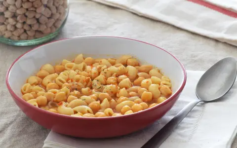 Un plato humeante de pasta e ceci, con garbanzos y macarrones en una salsa rojiza, junto a un tarro de garbanzos secos.