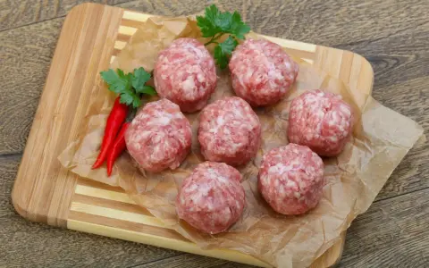 Surowe kotlety mielone keto, przyprawione papryczkami chili i natką pietruszki, leżą na papierze do pieczenia na desce.