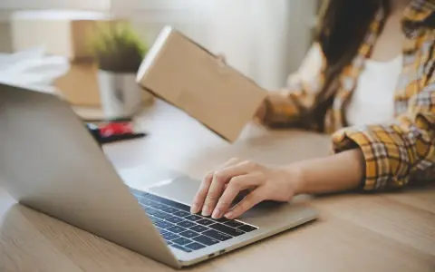Osoba pakuje paczkę, sprawdzając na laptopie, kto ponosi koszty zwrotu towaru.