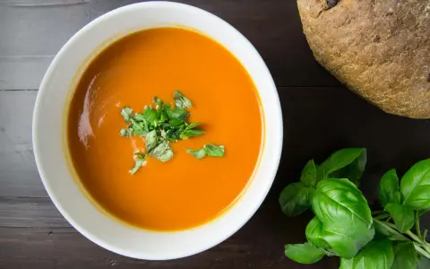 Pomidorowa zupa w białej misce, udekorowana bazylią, obok bochenek chleba.