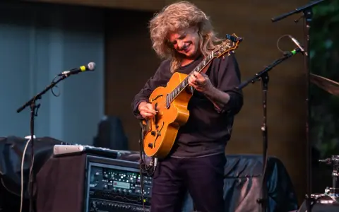 Muzyk z burzą blond włosów gra na gitarze, tworząc wspaniały śpiew w duecie z niewidocznym wokalistą.