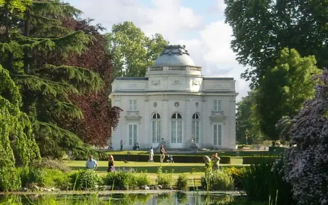 Un elegante edificio blanco con cúpula se alza en un exuberante jardin des plantes, con un estanque en primer plano y gente paseando.