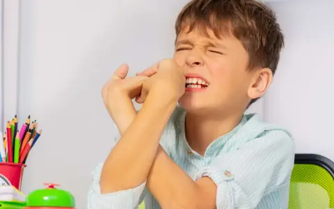 Junge mit zusammengepressten Augen und Händen vor dem Gesicht, ein Zeichen dafür, wie mit Tics bei Kindern umgehen. Buntstifte und eine Klingel liegen auf dem Tisch.