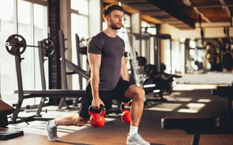 Mężczyzna wykonuje wykroki z kettlebellami, realizując swój plan treningowy na siłownię.