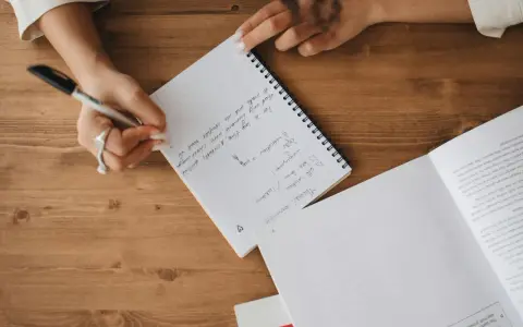 A person's hands are writing notes in a notebook. The notes mention "Theme: coronavirus", "self-isolation", and "lockdown".