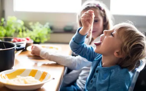 Zwei Kinder am Tisch, die schnell Nudeln essen. Der Junge nimmt eine Gabel voll Spaghetti zum Mund.