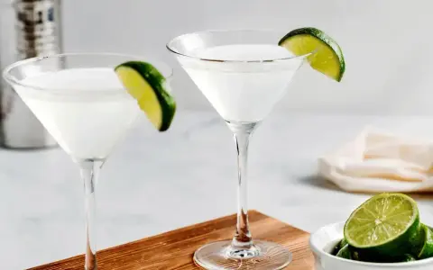 Dwa kieliszki z białym drinkiem, ozdobione plasterkami limonki. Obok miseczka z limonkami i shaker. Idealny kamikaze shot na gorący wieczór.
