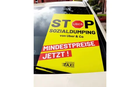 Gelbes Schild auf Taxidach: "STOP SOCIAL DUMPING von Uber & Co. MINDESTPREISE JETZT!". Fordert faire taksi fiyatları.