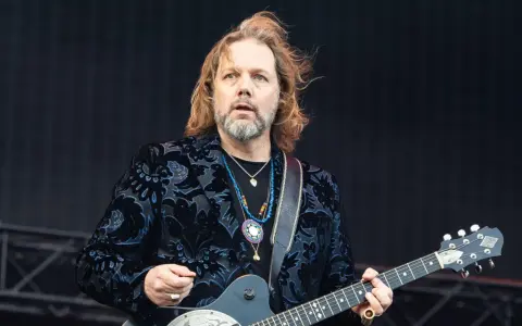 Rich Robinson, guitarrista con cabello largo y barba, luce un saco negro con estampado floral azul y varias cadenas.