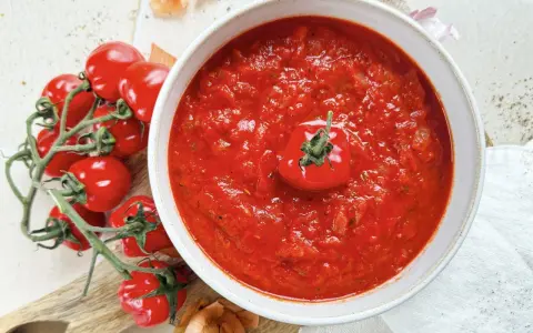 Ein Teller mit leuchtend rotem Sugo di Pomodoro, garniert mit einer einzelnen Kirschtomate. Daneben ein Bund frischer Tomaten und Zwiebeln.