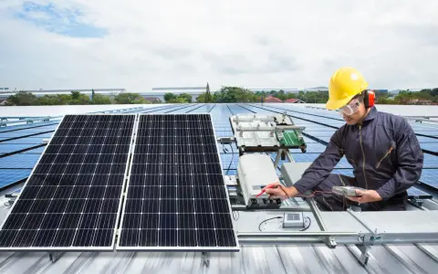 Technik w żółtym kasku sprawdza instalację solar edge na dachu. Panele słoneczne błyszczą w słońcu.