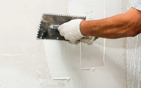 Mano enguantada aplicando estuco a una pared con una llana dentada. El proceso de estucar una pared requiere precisión.