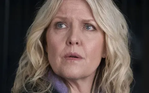 En kvinna med blont hår och blå ögon, klädd i lila, ser oroligt ut. Kanske är hon mitt i ett dramatiskt ögonblick i **shetland serie**.