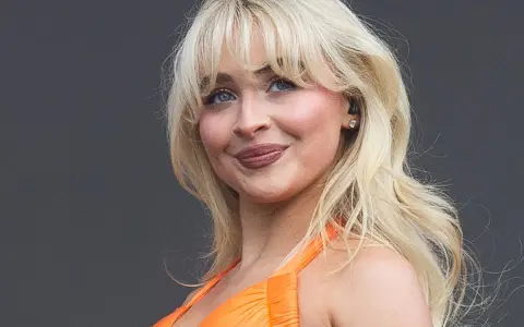Blonde Frau mit voluminösen Blow-out-Haaren und einem strahlenden Lächeln in einem orangefarbenen Kleid.