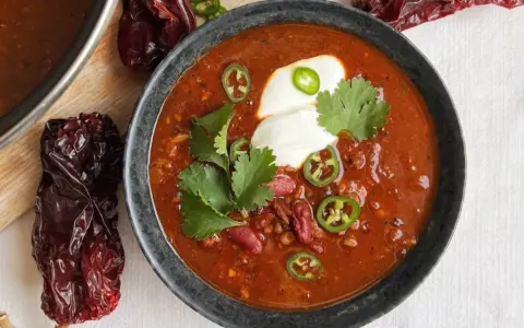 Un tazón humeante de chili con carne, adornado con crema agria, jalapeños y cilantro. Alrededor, chiles secos añaden un toque rústico.