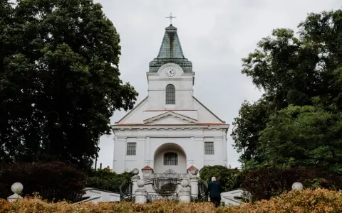 Biały kościół z zegarem na wieży, otoczony drzewami. Zastanawiasz się, jak sprawdzić do której parafii należę?