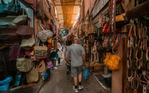 Bunte Taschen und Gürtel hängen in einem engen Gang des Marrakesch Basars. Ein Mann geht mit einer blauen Tüte durch das bunte Treiben.