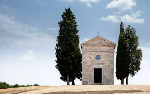 La cappella della Madonna di Vitaleta, con i suoi cipressi iconici, si erge solitaria su una collina toscana sotto un cielo azzurro e nuvoloso.